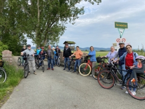 Gruppe von Menschen auf einer Straße in Landschaft vor einer steinernen Brücke, alle angelehnt an ihren Fahrrädern.